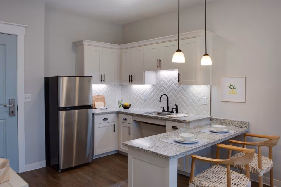 Modern kitchen with white cabinets, granite countertops, a stainless steel refrigerator, a black faucet, and two pendant lights hanging over a breakfast bar with two wooden chairs set with white bowls and plates.