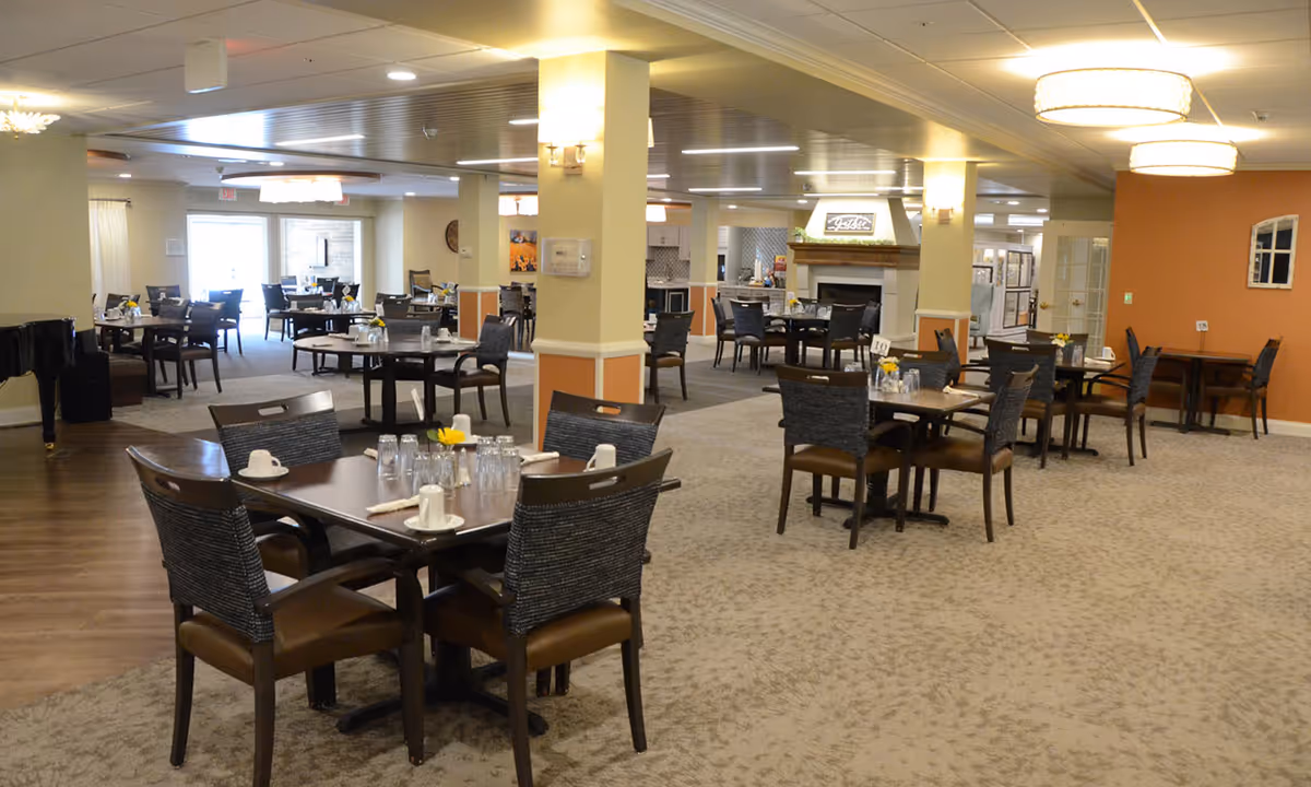 Spacious senior living dining room with multiple set tables and chairs, warm lighting, and a fireplace in the background.