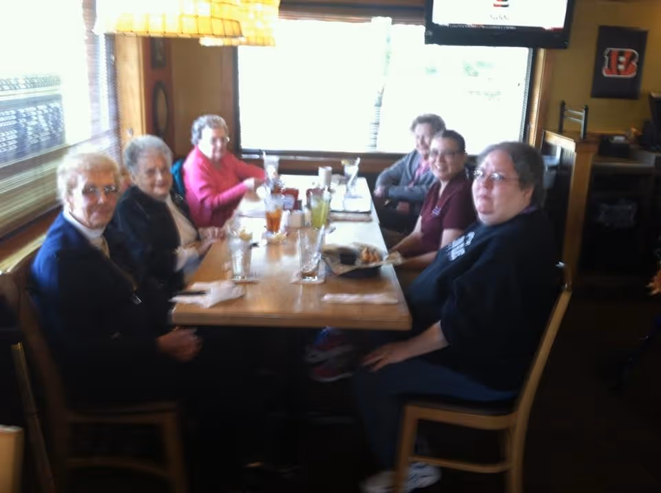 A group of six people sitting around a wooden dining table in a restaurant, with drinks and plates in front of them. The setting is cozy with large windows and warm lighting.