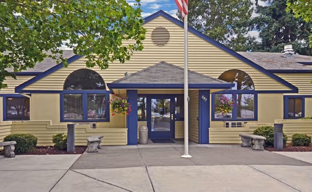 Front exterior view of a single-story building with yellow siding and blue trim, featuring a central entrance with a covered porch, an American flag on a flagpole in front, hanging flower baskets on either side of the entrance, and concrete benches along the walkway.