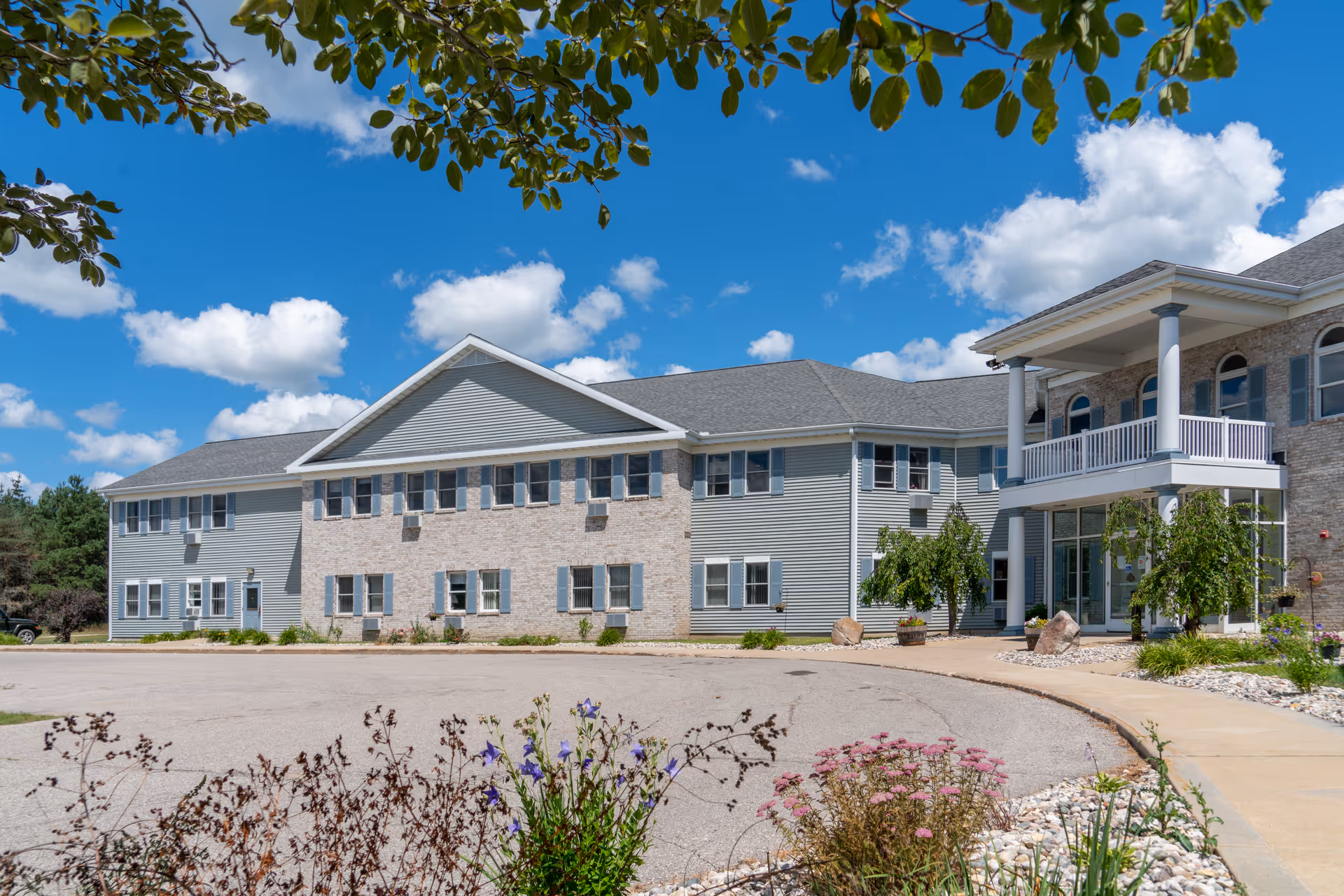 Exterior view of a senior living facility building under a bright blue sky with scattered clouds. The building has light gray siding with stone accents, multiple windows with blue shutters, and a covered entrance with white columns and a balcony. There are landscaped plants and flowers in the foreground along a curved driveway.