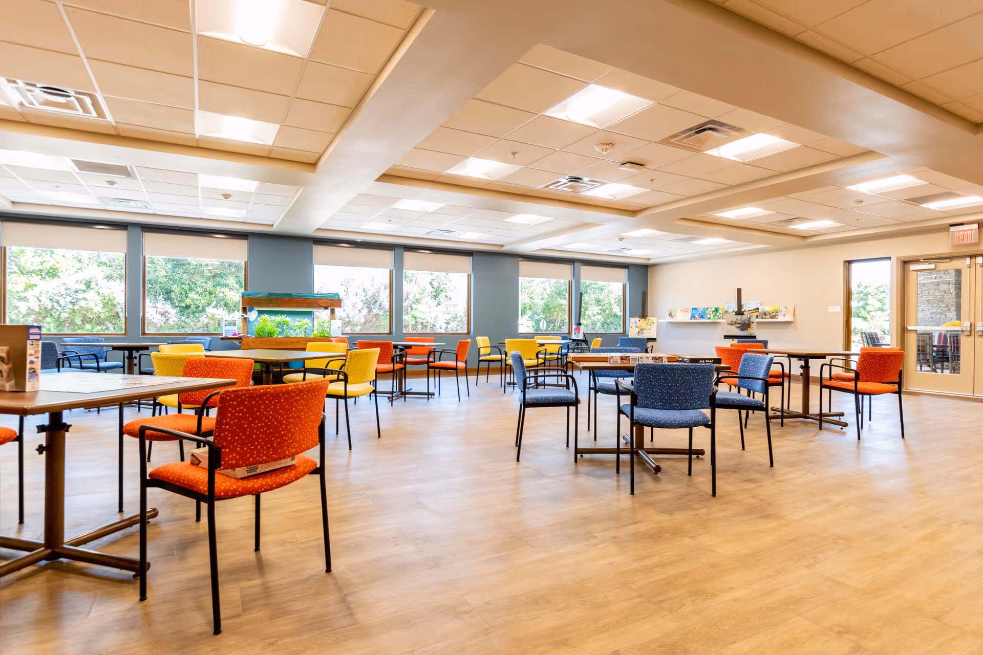 Spacious, brightly lit communal dining/activity room with multiple tables and colorful chairs and large windows.