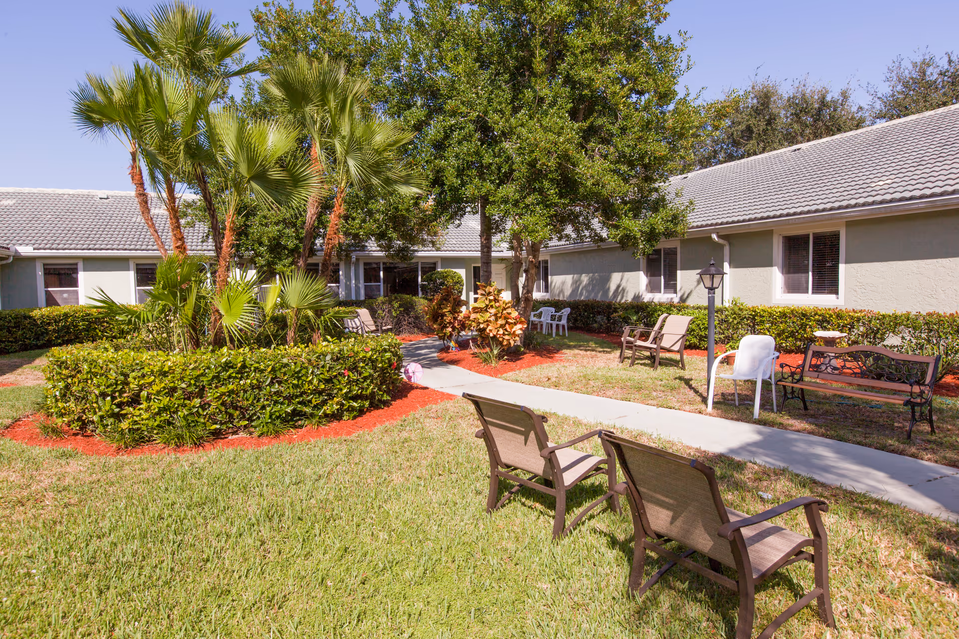 Outdoor courtyard area at Brookdale Tequesta featuring a variety of seating options including benches and chairs arranged along a paved walkway. The space is landscaped with green grass, trimmed bushes, palm trees, and other plants under a clear blue sky.