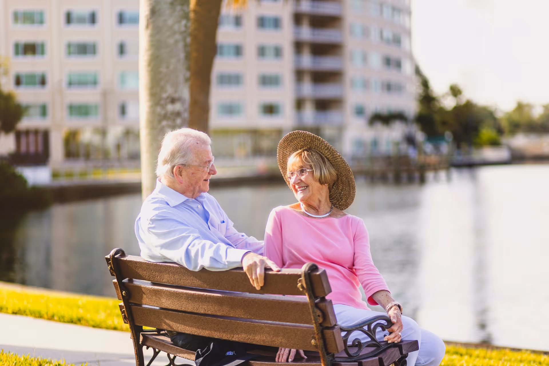 An elderly man and woman sitting on a wooden bench by a waterfront, smiling and engaging in conversation. The woman is wearing a pink top and a wide-brimmed straw hat, while the man is dressed in a light blue shirt. Behind them is a tree and a large building across the water.