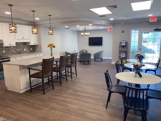 A spacious and well-lit common area featuring a kitchen with white cabinets, a large island with bar stools, and pendant lights. There are round tables with chairs and flower centerpieces, a seating area with two armchairs facing a wall-mounted TV, and large windows and doors letting in natural light.