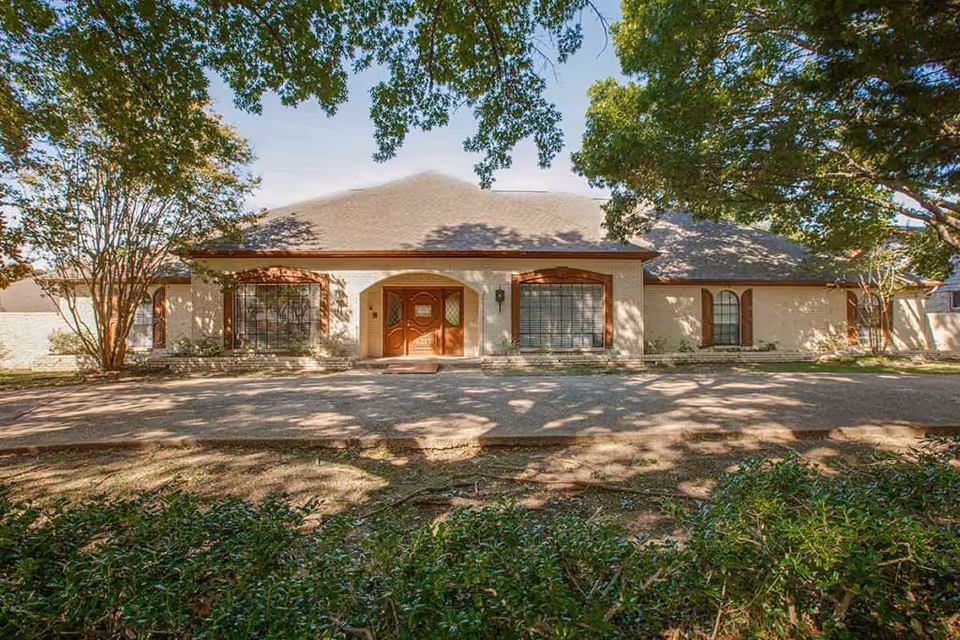 Front exterior view of a single-story building with a sloped roof, large windows with decorative shutters, and a central wooden double door entrance. The building is surrounded by trees and greenery, with shadows cast on the driveway and lawn in front.