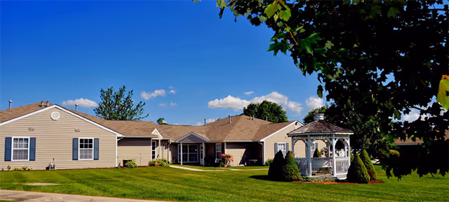 Single-story beige residential building with multiple sections and a brown shingled roof under a clear blue sky. In front of the building is a well-maintained green lawn with a white gazebo surrounded by shrubs and trees.