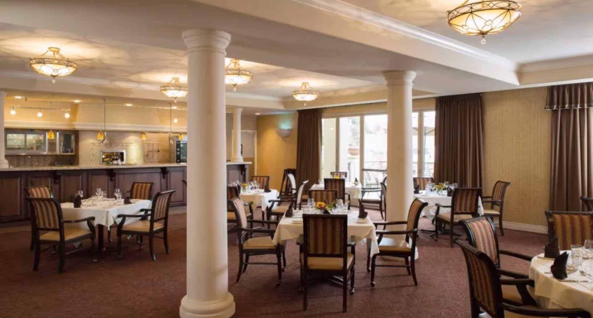 Dining room with round tables draped in white linens, wooden chairs, decorative columns, and a serving counter under warm lighting.