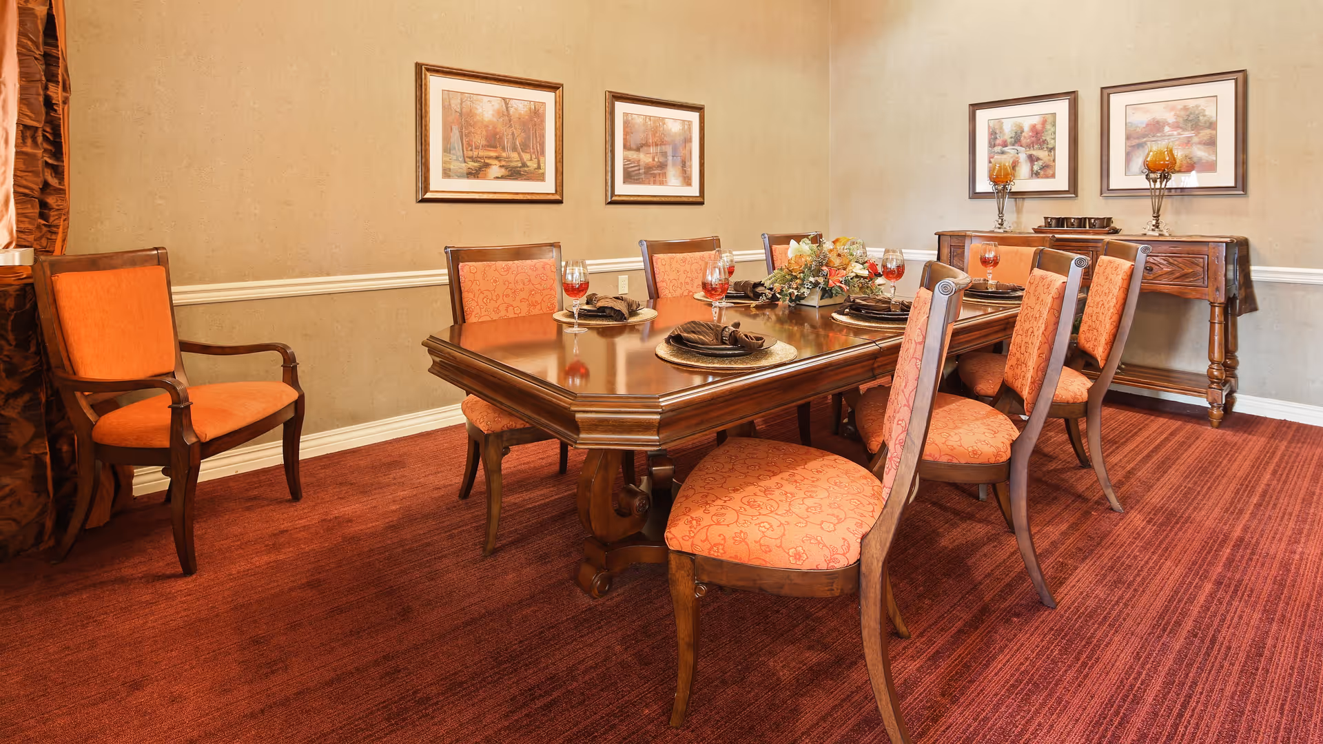 A formal dining room with a wooden dining table set for six people. The chairs have orange upholstered seats and backs. The table is decorated with a floral centerpiece and place settings with glasses of red beverage. The room has beige walls adorned with framed landscape paintings and a sideboard with decorative candle holders.