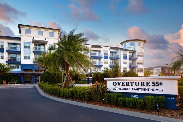Exterior view of Overture 55+ Active Adult Apartment Homes building with palm trees and landscaped greenery under a partly cloudy sky during sunset.