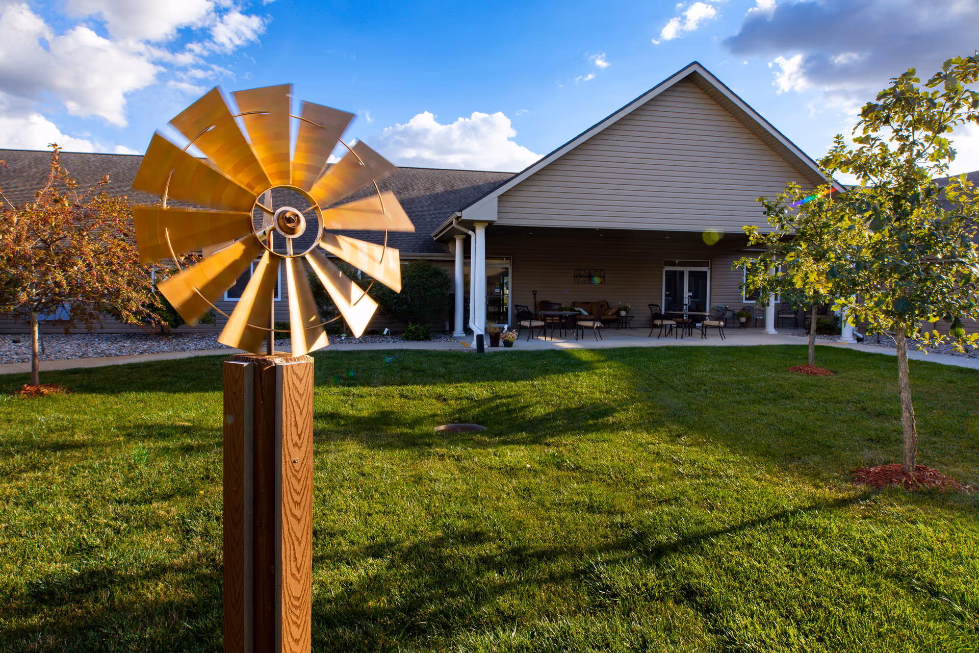 A landscaped lawn and patio in front of a single-story assisted living building with a decorative metal windmill sculpture in the foreground.