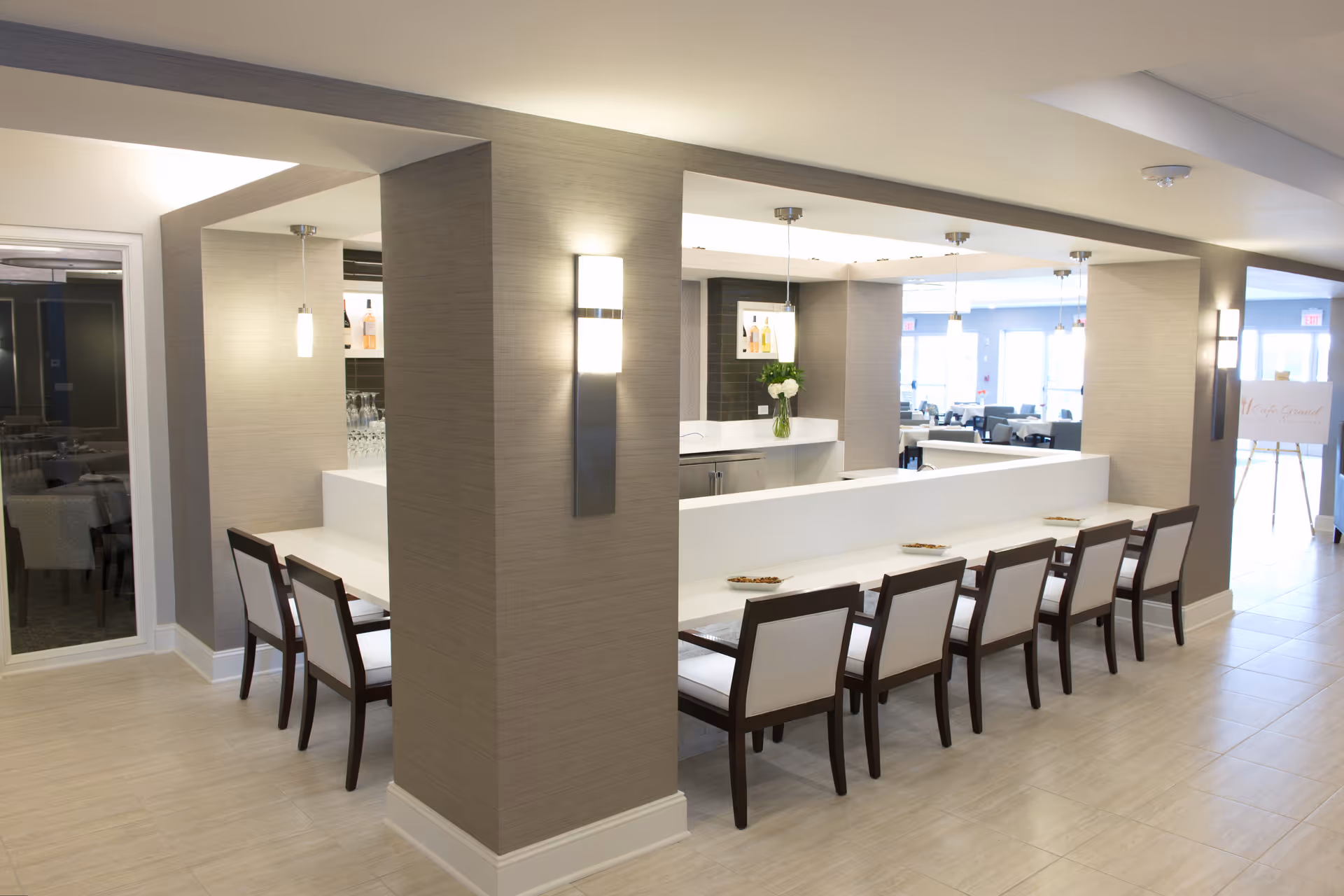 A modern dining area with a long white counter surrounded by eight chairs with dark wooden frames and white cushions. The space is well-lit with wall sconces and pendant lights hanging from the ceiling. In the background, there is a bar area with shelves holding bottles and glassware, and large windows letting in natural light.