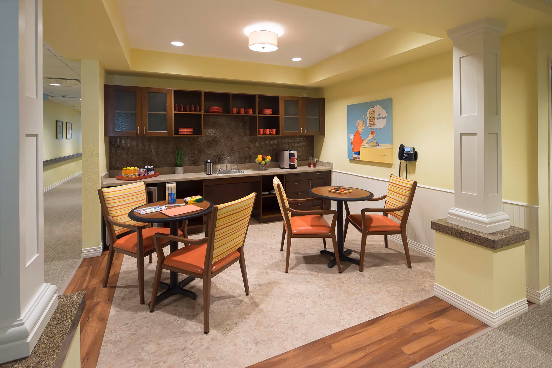 A small dining area in a senior living facility with two round tables, each surrounded by two chairs with orange cushions and striped backs. The room has a kitchenette with dark wood cabinets, a countertop with a sink, a coffee maker, and shelves holding red cups and plates. The walls are painted yellow, and there is a colorful painting on the wall. The floor is a combination of wood and tile, and the area is well-lit with ceiling lights.