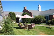 Outdoor courtyard area at Webster at Rye featuring a green lawn with a round table and four cushioned chairs under a pink umbrella. The courtyard is surrounded by a brick building with white trim and a small cupola on the roof. There are bushes and trees around the lawn under a clear blue sky.