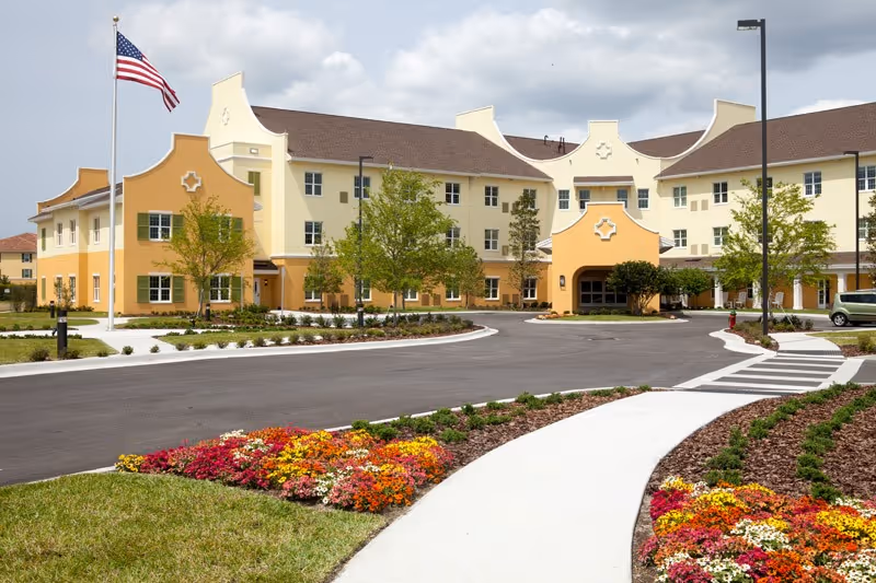 Exterior view of Discovery Village Trinity, a large senior living facility with a curved driveway, landscaped flower beds, green lawns, and an American flag on a flagpole in front of the building under a partly cloudy sky.