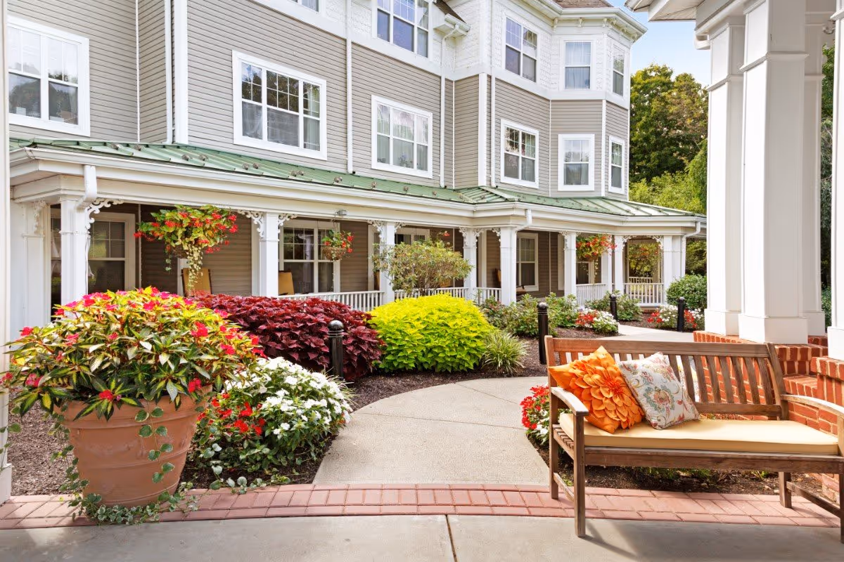 Outdoor seating area at Benchmark at Stamford featuring a wooden bench with colorful cushions, surrounded by lush greenery and flowering plants. The building exterior has beige siding with white trim and multiple windows, and a covered porch with decorative columns.