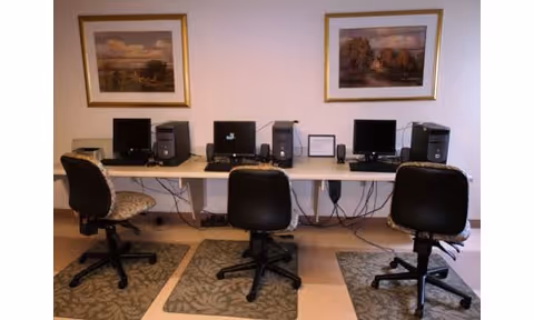 A computer workstation area with three desktop computers on a long white table. Each computer has a monitor, keyboard, and mouse. Three office chairs are positioned in front of the computers. Two framed landscape paintings hang on the wall above the table. The floor has patterned mats under each chair.