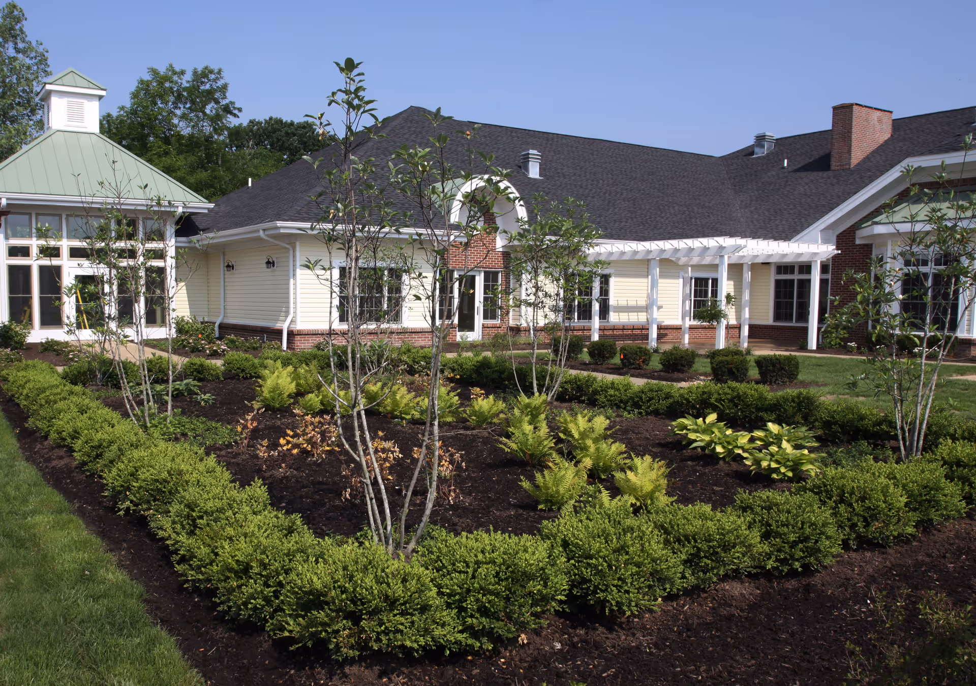 Exterior view of Deupree House showing a well-maintained garden with various shrubs and small trees in front of a building with light yellow siding, white trim, and a dark roof under a clear blue sky.
