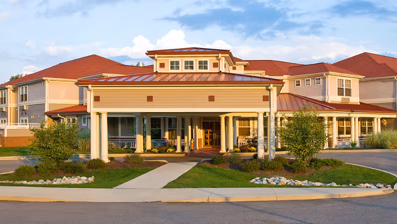 Front exterior view of a senior living facility building with a covered entrance supported by white columns, landscaped greenery, and a red roof under a partly cloudy sky.
