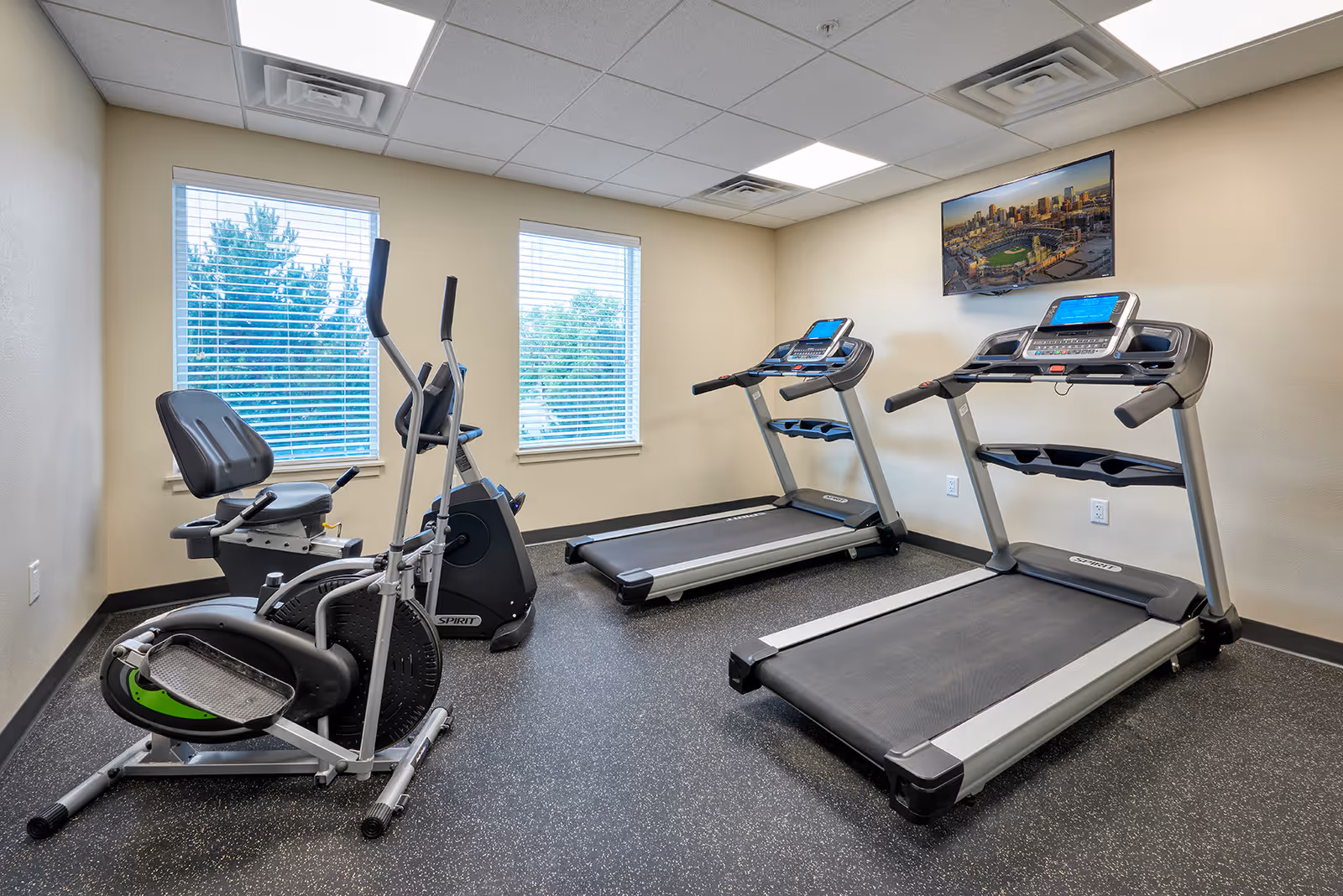 A small fitness room with two treadmills, an elliptical machine, and a recumbent bike. The room has two windows with blinds, beige walls, and a TV mounted on the wall displaying a cityscape with a baseball stadium.