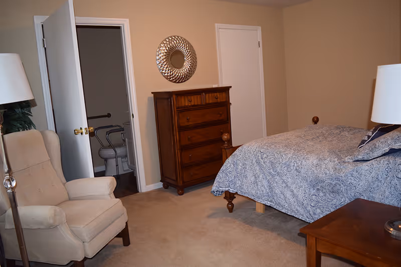 A bedroom with a wooden bed covered with a blue patterned bedspread, a wooden chest of drawers with a decorative round mirror above it, a beige upholstered armchair, a wooden side table with a lamp, and an open door leading to a bathroom with a toilet and grab bars.