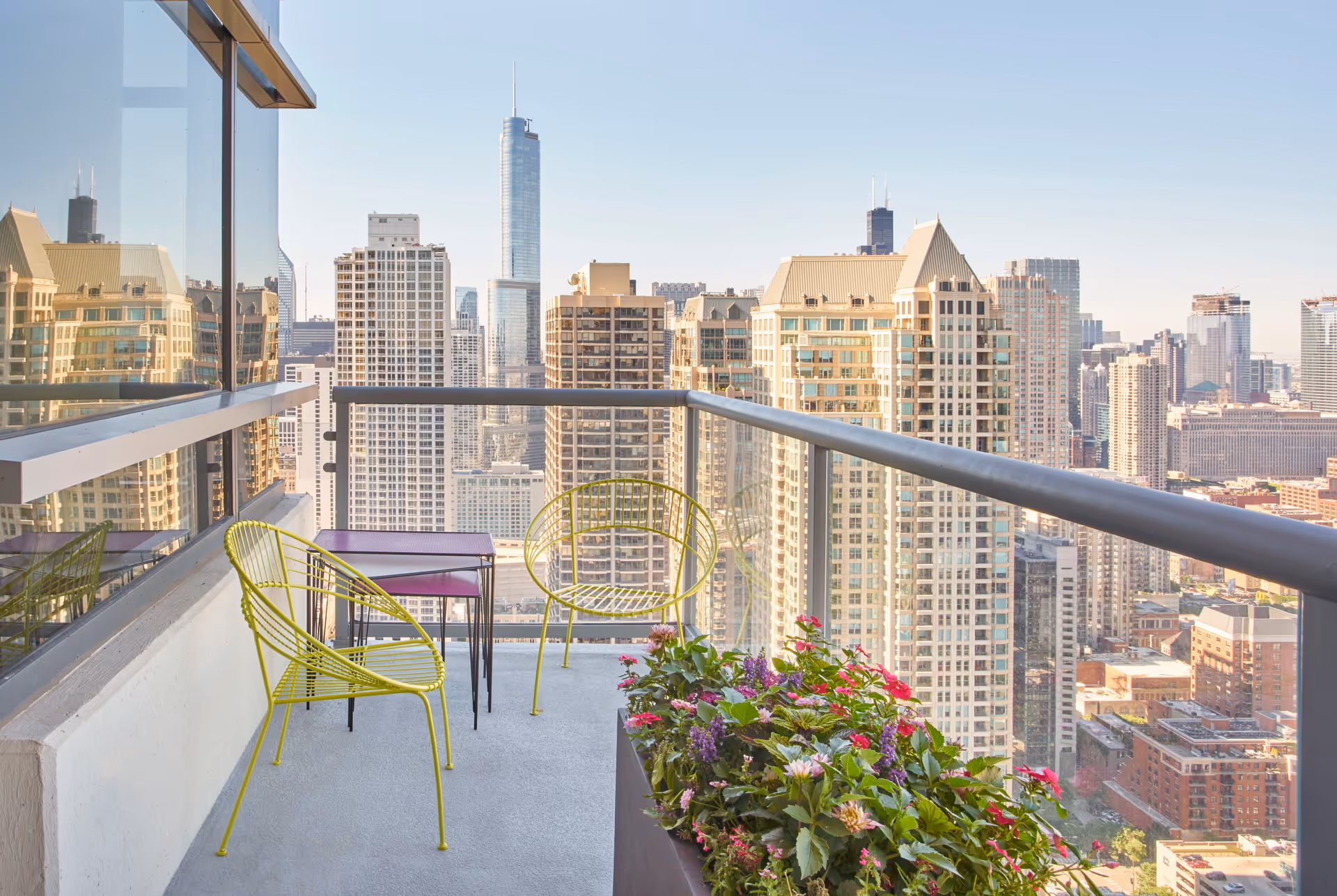 Balcony with two yellow metal chairs and a small purple table, overlooking a city skyline with tall buildings under a clear sky. A planter with colorful flowers is on the right side of the balcony.