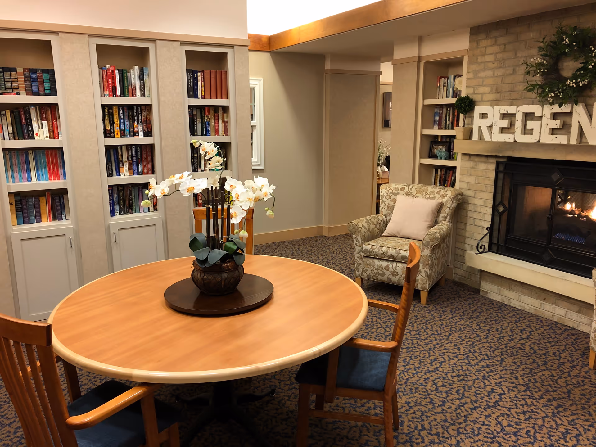Cozy senior living community room with a round wooden table and chairs, a potted orchid centerpiece, built-in bookshelves filled with books, a comfortable armchair with a pillow, and a lit fireplace with decorative letters spelling 'REGEN' on the mantel.