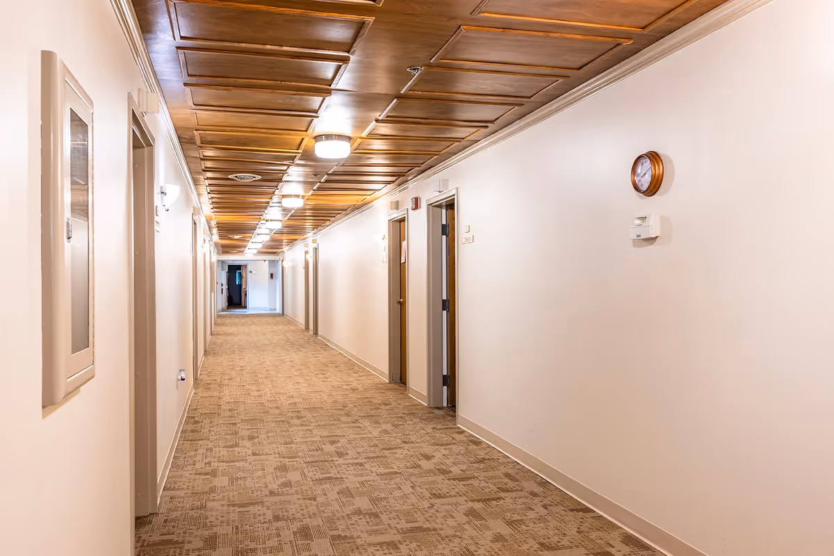 A long, well-lit hallway in an assisted living facility with beige carpet, white walls, wooden paneled ceiling, several doors along the sides, a wall clock, and a thermostat.