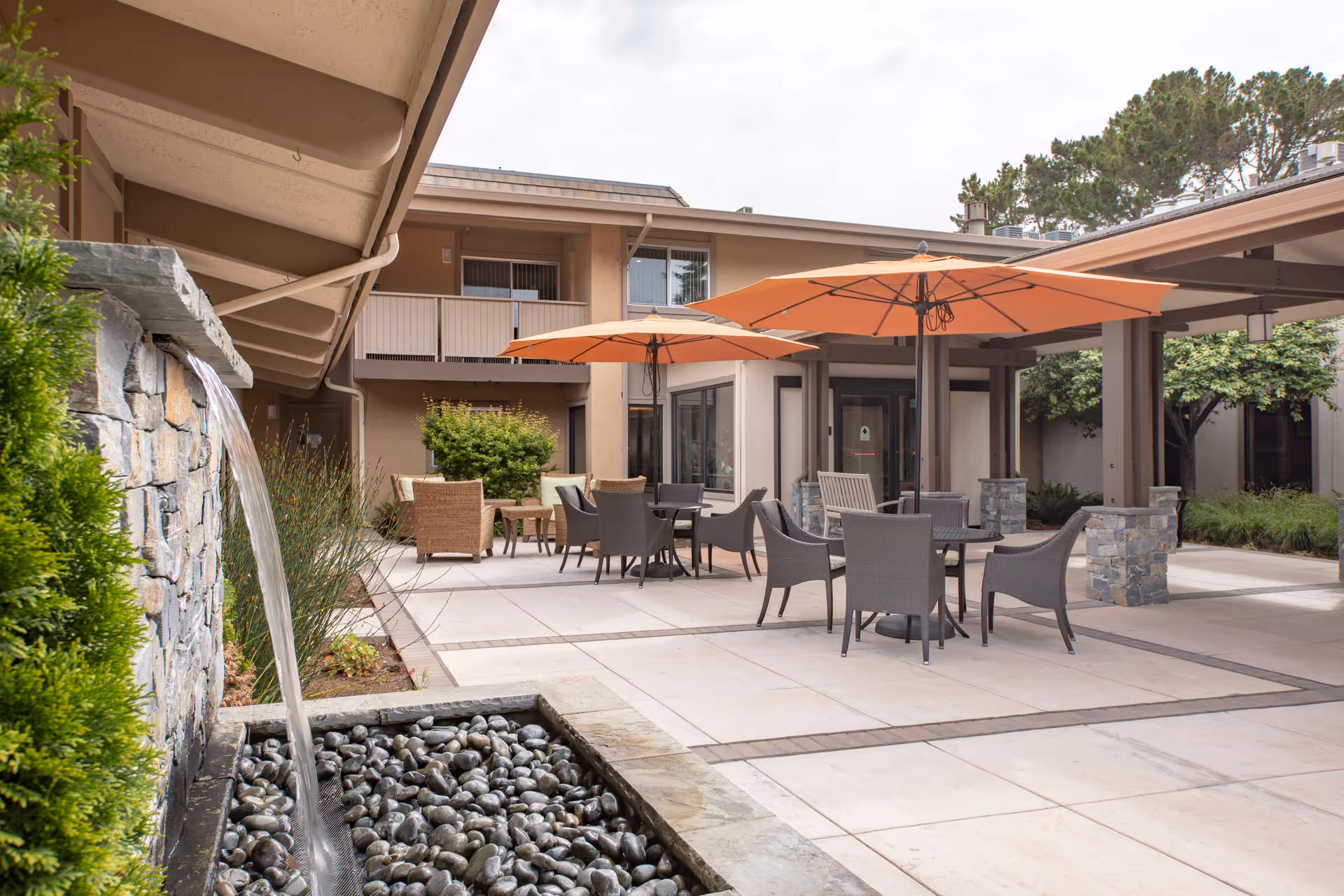 Outdoor courtyard area at BridgePoint at Los Altos featuring a stone water fountain with flowing water, several tables with chairs, and large orange umbrellas providing shade. The courtyard is surrounded by a two-story building with windows and greenery.