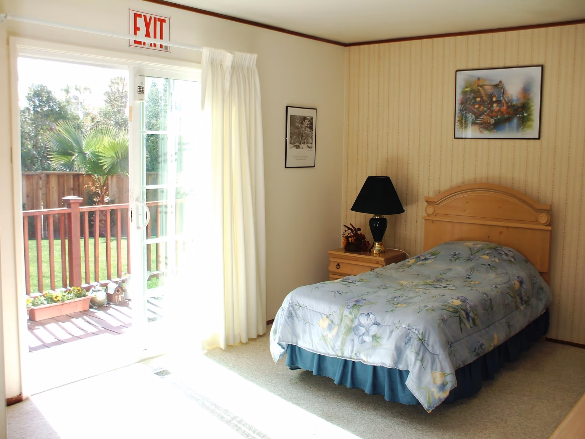 A bright bedroom with a single bed covered in a floral comforter, a wooden nightstand with a black lamp and a small flower arrangement, two framed pictures on the walls, and a sliding glass door leading to an outdoor deck with a wooden railing and greenery visible outside.