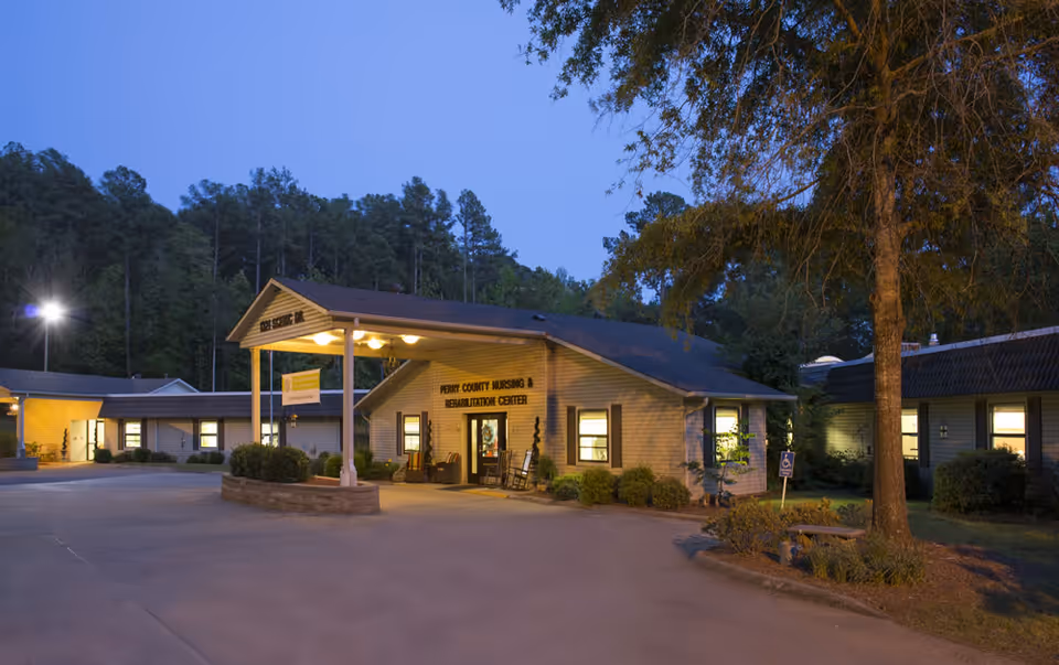 Exterior view of Perry County Nursing & Rehab Center at dusk, showing a single-story building with a covered entrance, illuminated windows, surrounding trees, and a paved driveway.