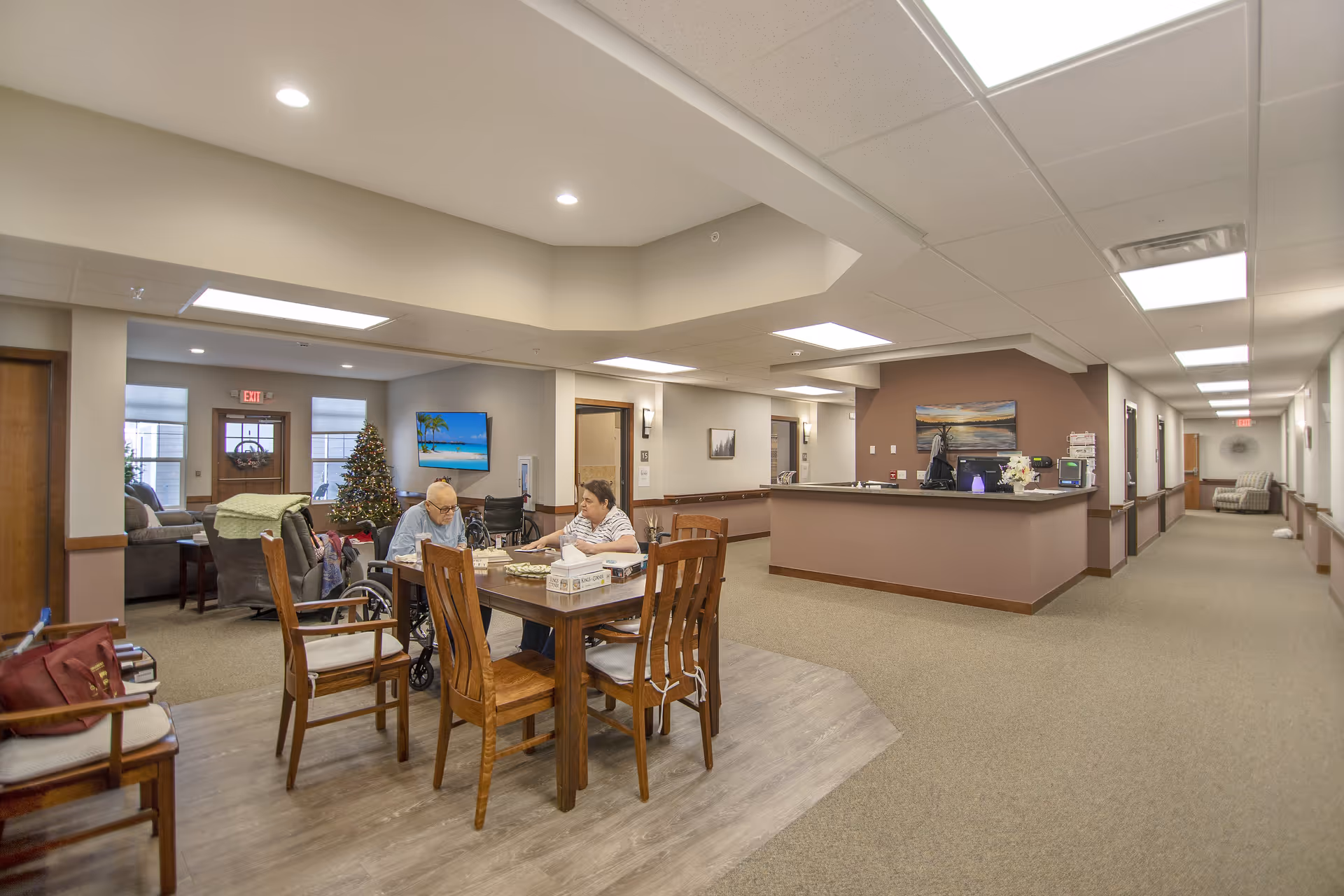 A senior living facility common area with two elderly individuals sitting at a wooden table playing a board game. The room has a Christmas tree, a television mounted on the wall displaying a beach scene, and a reception desk. The area is well-lit with ceiling lights and has a hallway leading to other rooms.
