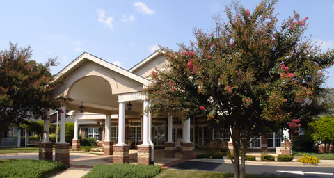 Front entrance of a senior living building with a covered porte-cochere, brick columns and flowering trees under a blue sky.