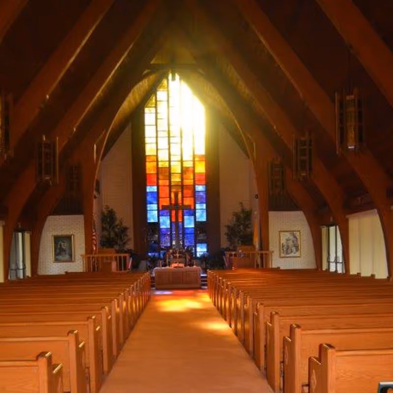 Interior of a chapel with wooden pews, arched wooden ceiling, and a large colorful stained-glass window above the altar.