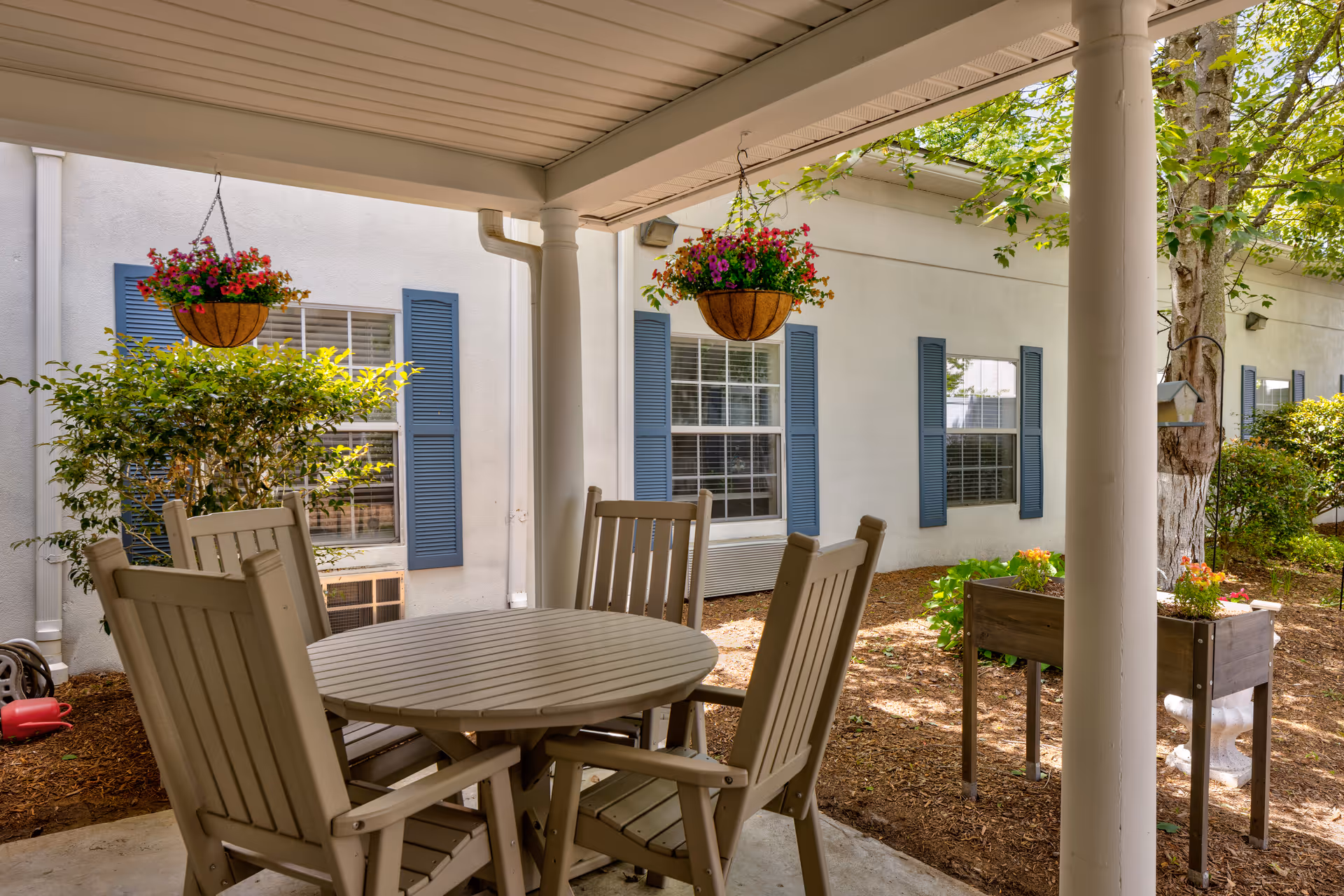 Covered patio with a round table and wooden chairs, hanging flower baskets, planter boxes, and a building exterior with windows and blue shutters.