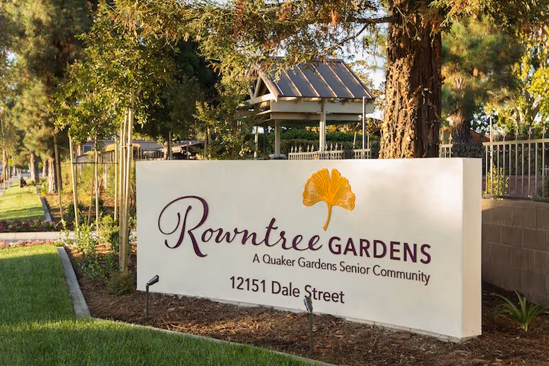 Outdoor view of a white sign for Rowntree Gardens, a Quaker Gardens Senior Community, located at 12151 Dale Street, surrounded by trees and greenery.