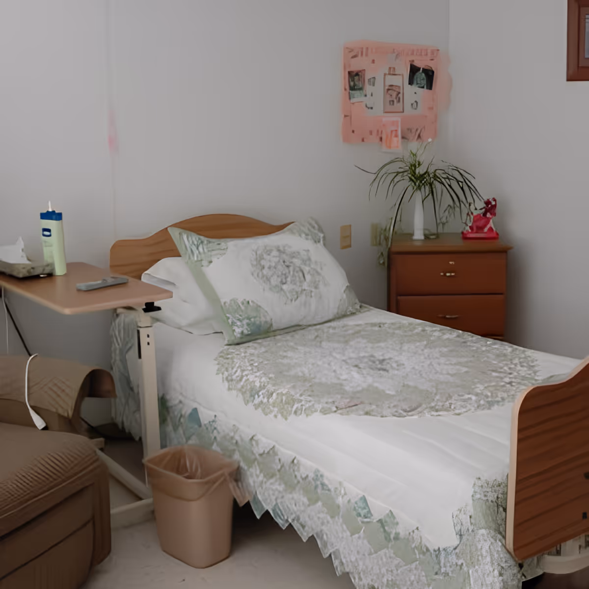 A single bed with a floral patterned bedspread and pillow in a small room. Next to the bed is a wooden nightstand with a plant and a red decorative item on top. There is a small table attached to the bed holding a lotion bottle and a remote control. A trash bin is placed on the floor beside the bed. The walls are plain white with a pink bulletin board displaying photos and notes.