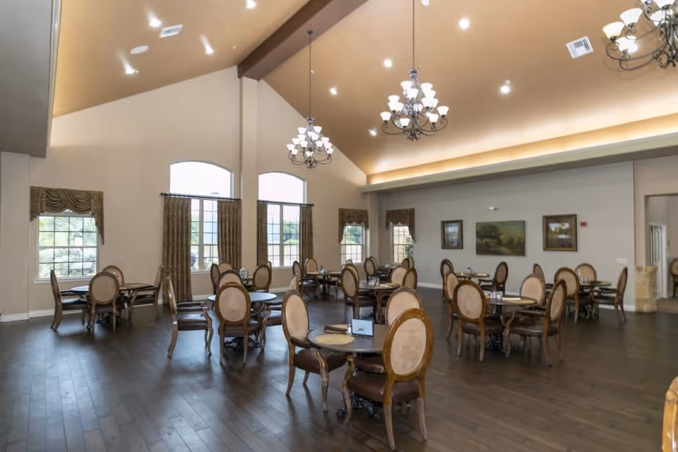 Spacious dining room with multiple round tables and cushioned wooden chairs arranged neatly. The room has high vaulted ceilings with elegant chandeliers and large windows with draped curtains allowing natural light to enter. The walls are decorated with framed paintings.