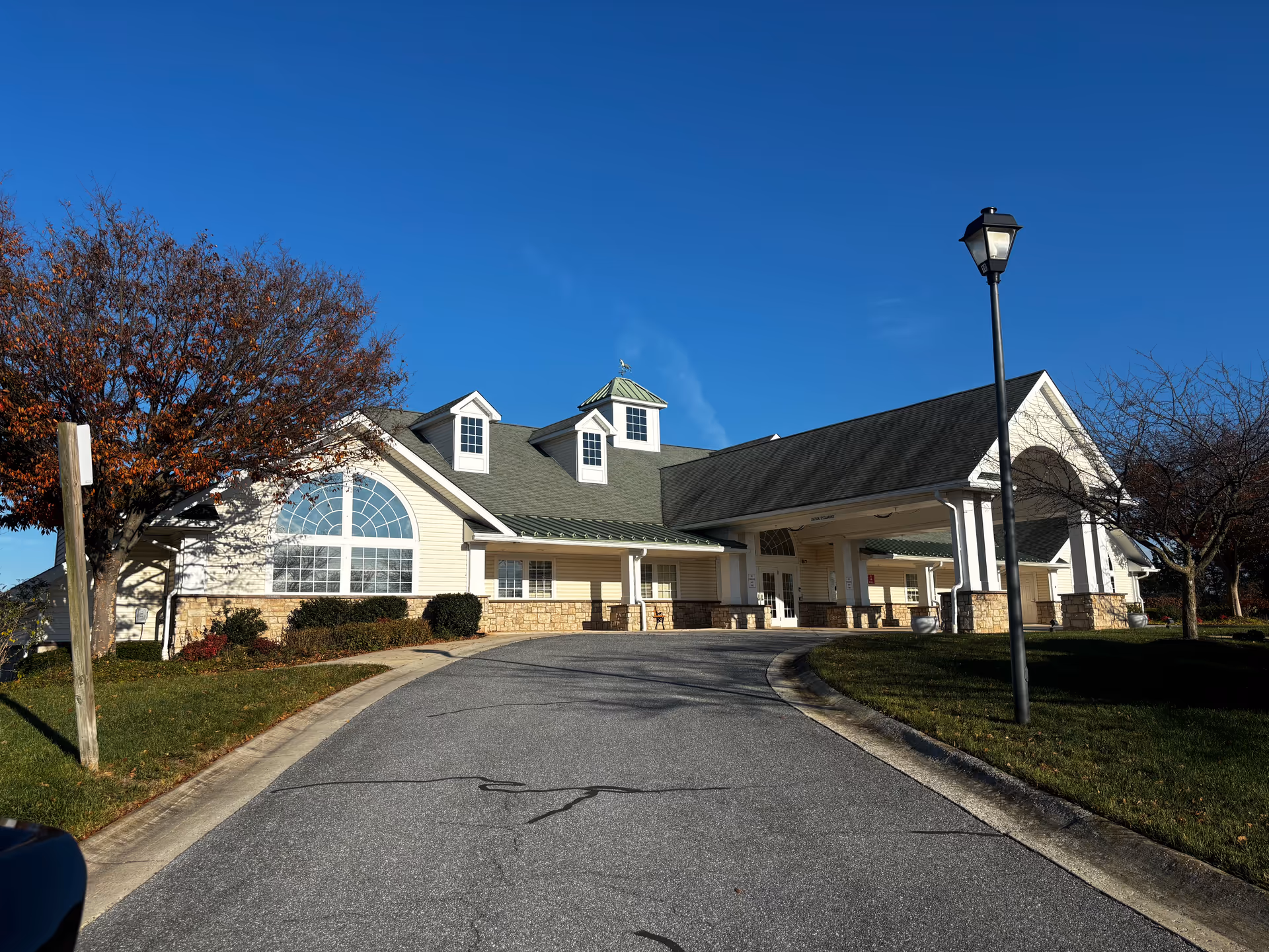 Exterior view of Carroll Vista facility showing a large building with a covered entrance, multiple windows including a large arched window, a green roof with dormer windows, a street lamp, and trees with autumn foliage under a clear blue sky.