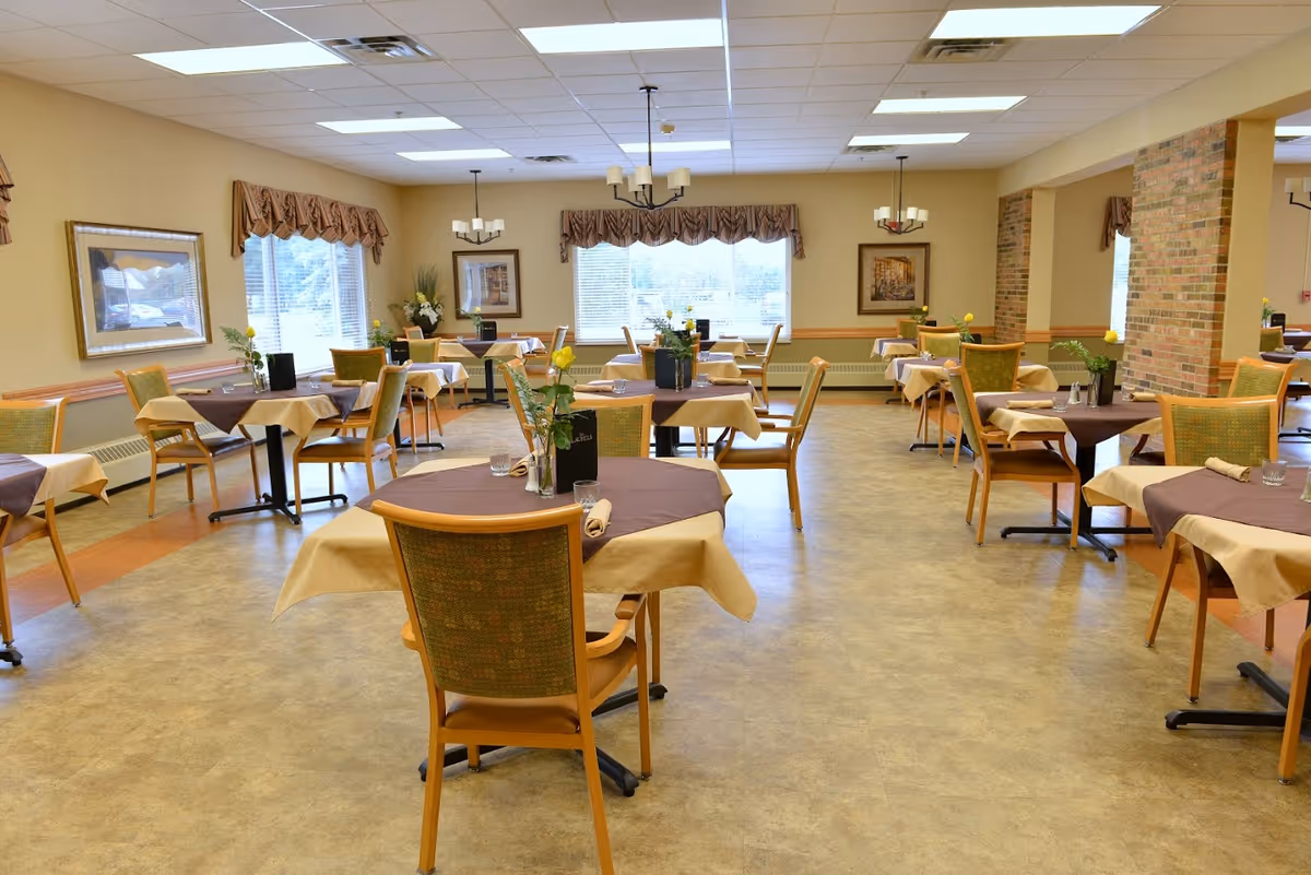 A spacious dining room with multiple tables covered with beige and brown tablecloths, each set with chairs, small flower vases, and menus. The room has large windows with valance curtains, framed artwork on the walls, and ceiling lights providing bright illumination.