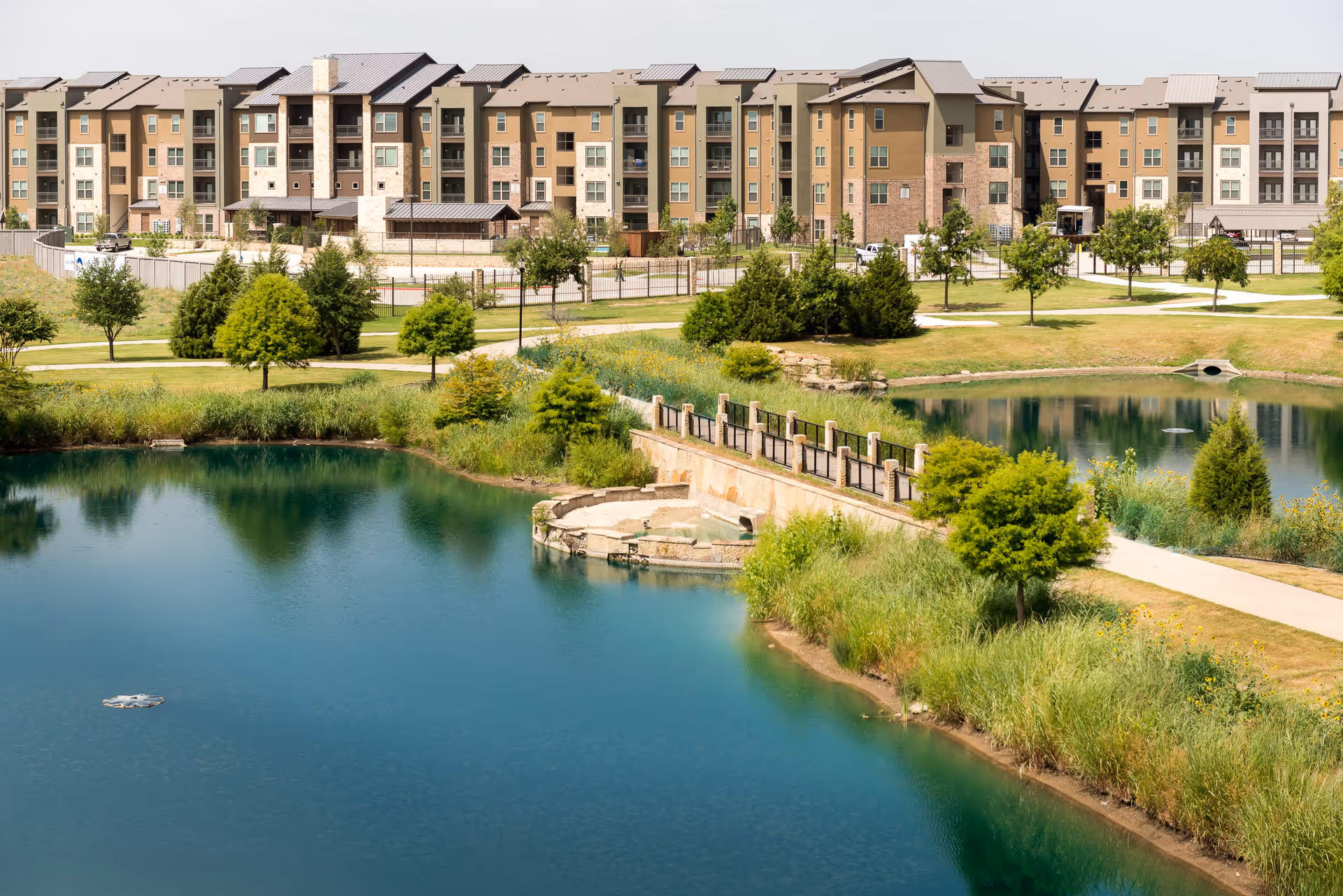 A multi-story senior living apartment building overlooks landscaped grounds with walking paths, trees, a bridge and a pond.