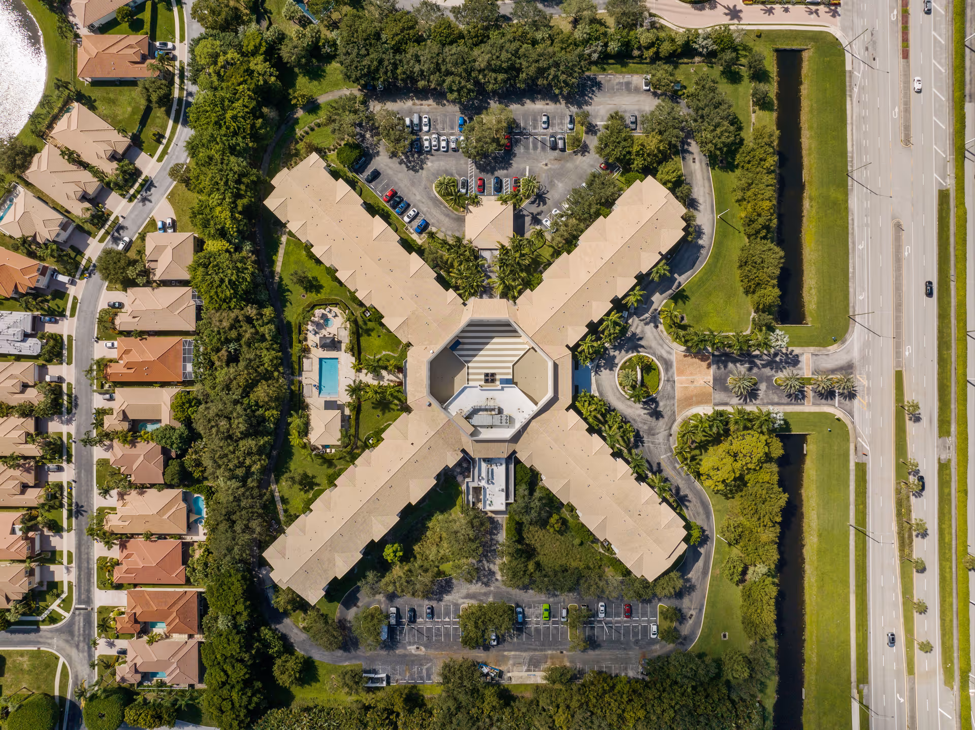 Aerial view of Verena at Delray facility showing a large X-shaped building surrounded by parking lots, trees, and nearby residential houses. Roads and green spaces are visible around the property.