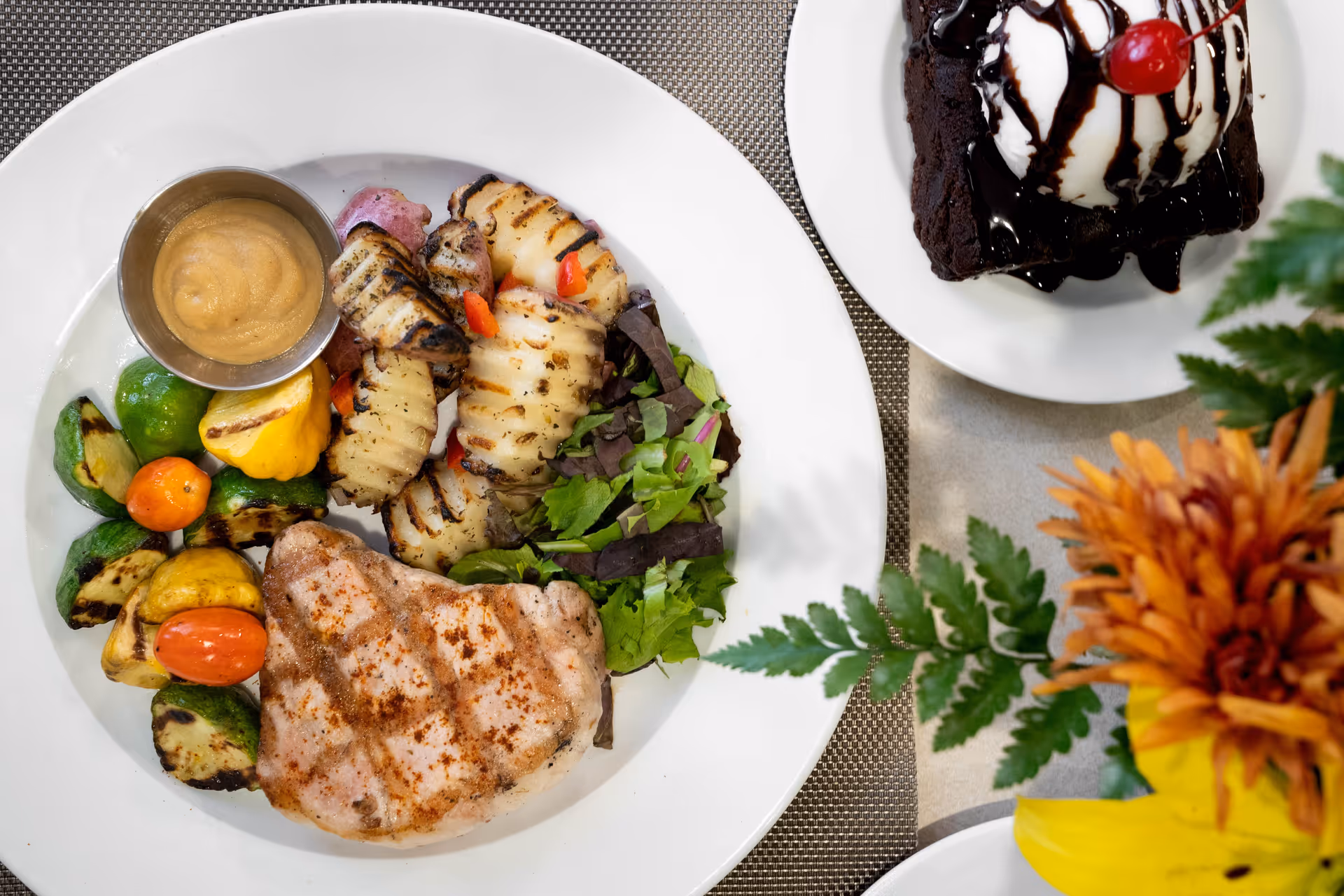 A plate of grilled food including a piece of grilled chicken, grilled vegetables, grilled squid, a small serving of sauce, and a side of mixed greens. Next to the plate is a dessert consisting of a brownie topped with vanilla ice cream, chocolate syrup, and a cherry. There is also a floral arrangement with orange and yellow flowers on the table.