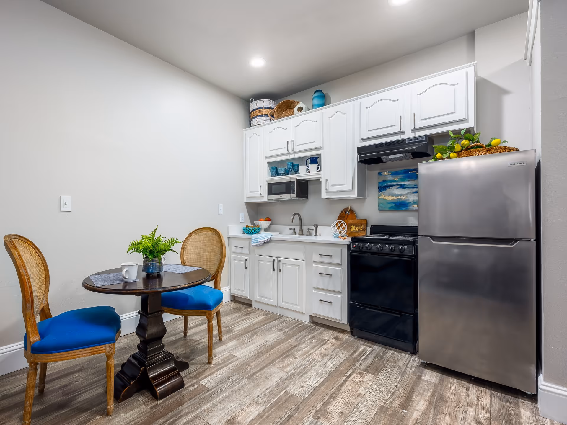 A small kitchen area with white cabinets, a stainless steel refrigerator, a black stove with an overhead vent, and a sink. There is a round wooden table with two chairs that have blue cushions. The floor is wood-patterned, and the walls are light-colored. Decorative items include a plant on the table, baskets on top of the cabinets, and a painting above the stove.