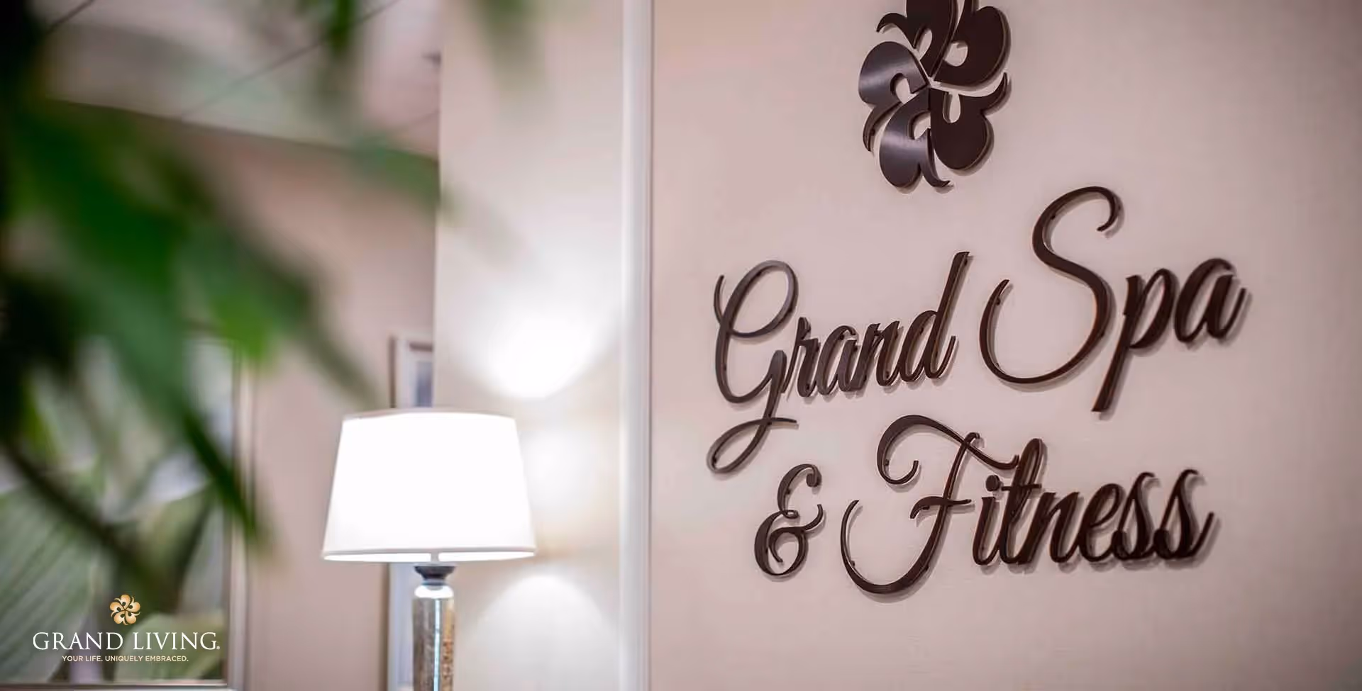 Interior wall with decorative text reading 'Grand Spa & Fitness' in elegant script, a lamp with a white shade, and some blurred green plant leaves in the foreground.