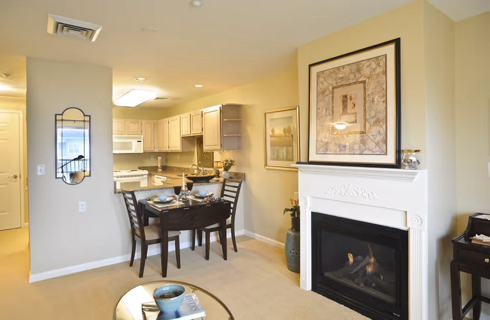 A cozy interior space featuring a small dining area with a dark wooden table set for four, adjacent to a kitchen with light wood cabinets and granite countertops. A white fireplace with decorative molding and a framed artwork above it is on the right side. The room has beige walls and carpet, with additional framed artwork and a decorative mirror on the walls.