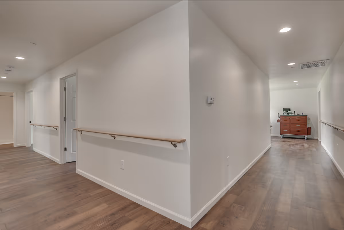 Bright hallway in a senior living facility with wood-like flooring and wall-mounted handrails.