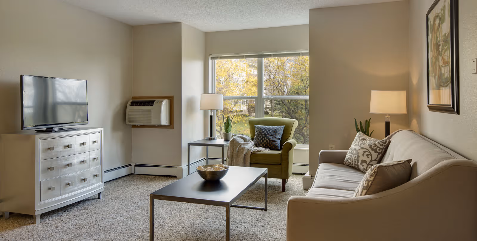 A cozy living room with a beige sofa adorned with patterned pillows, a green armchair with a throw blanket and a decorative pillow, a modern coffee table with a bowl of decorative items, a side table with a lamp and a small plant, a flat-screen TV on a white dresser, and a large window showing trees outside.