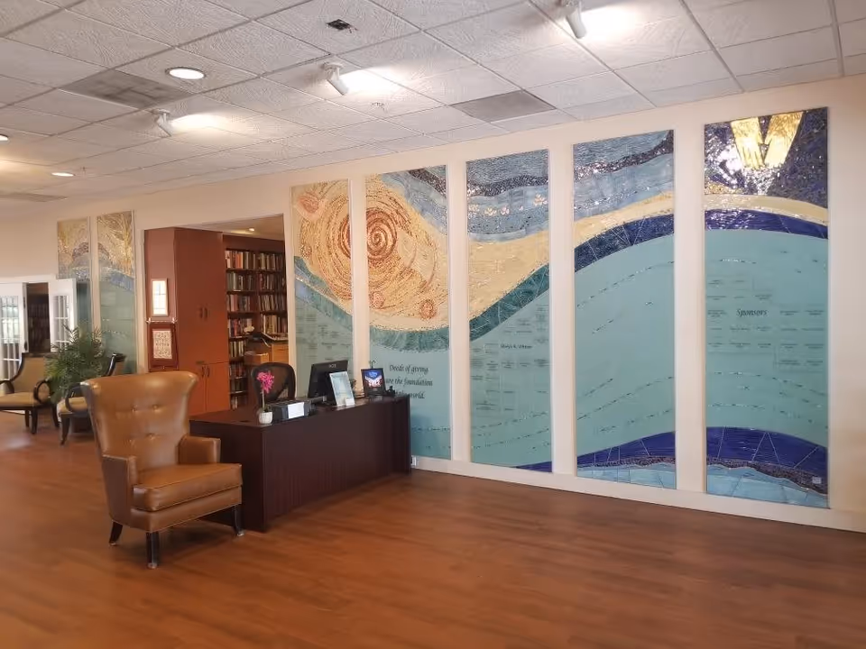 A reception area inside the Toby Weinman Residence featuring a wooden desk with a computer and a small potted plant. Behind the desk is a large decorative wall art composed of multiple panels with abstract designs in blue, beige, and brown tones. To the left, there is a brown leather armchair and a glimpse of a seating area with chairs and a bookshelf filled with books. The floor is wooden, and the ceiling has recessed lighting and white tiles.