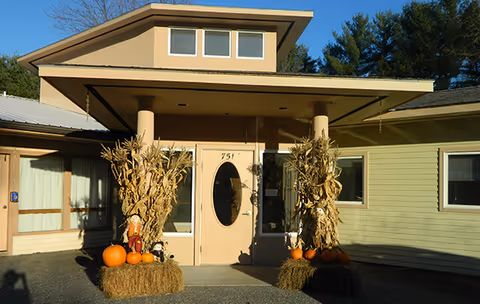 Entrance of The Sarah Frye Home decorated with autumn-themed items including hay bales, pumpkins, dried corn stalks, and scarecrows under a covered porch with beige walls and windows.